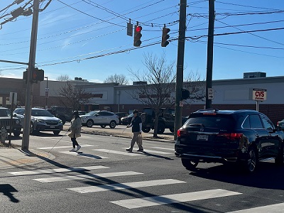 Student crosses street with cane as O&M instructor follows behind.